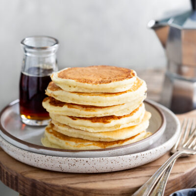 A stack of pancakes next to a bottle of syrup and small bowls of blueberries and chocolate chips.