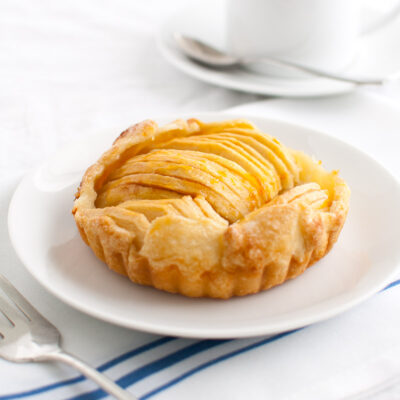 Apple tartlet on a white plate with a cup of coffee in the background.