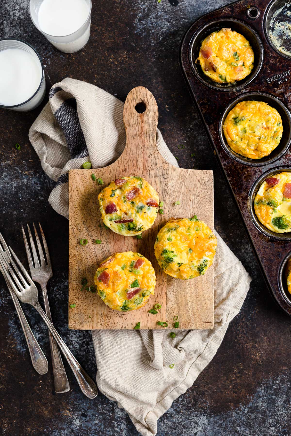 Three mini frittatas on a wooden board next to a muffin tin with more mini frittatas.