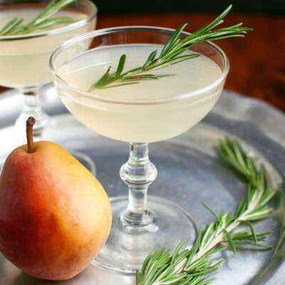 A pear-rosemary cocktail in a coupe glass garnished with a sprig of rosemary.