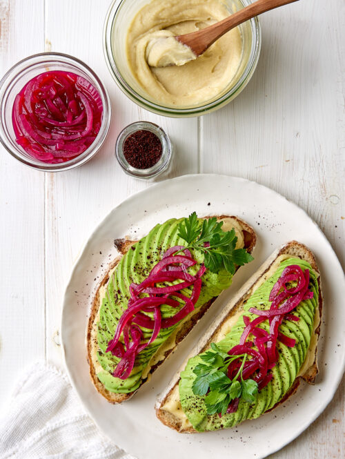 Two slices of avocado toast on a white plate next to some cashew cream and pickled onions.