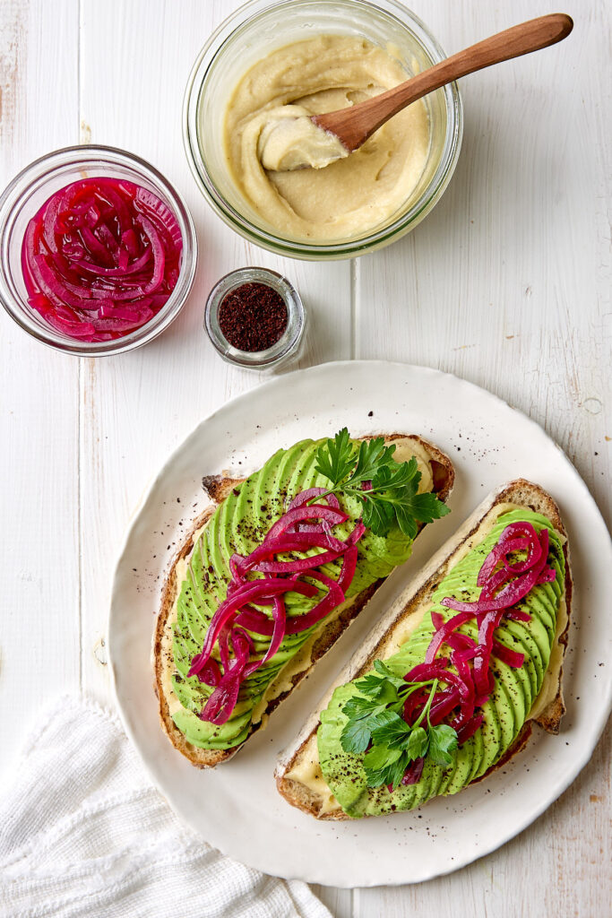 Two slices of avocado toast on a white plate next to some cashew cream and pickled onions.