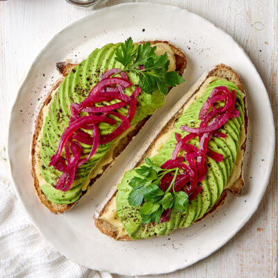 Two slices of avocado toast on a white plate next to some cashew cream and pickled onions.