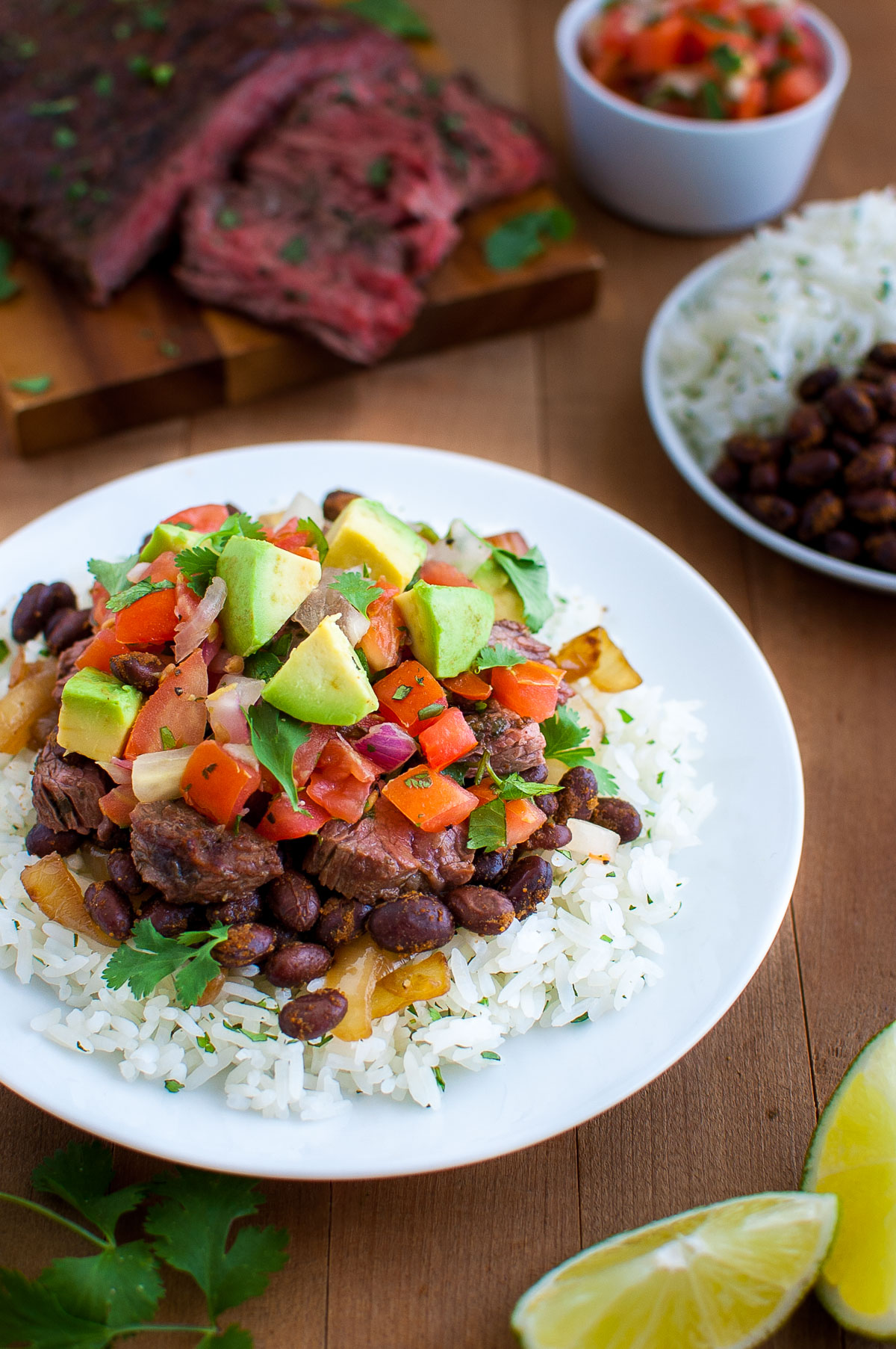 A plate of rice, black beans, chopped avocados, pico de gallo, and cut steak with more sliced steak in the background.