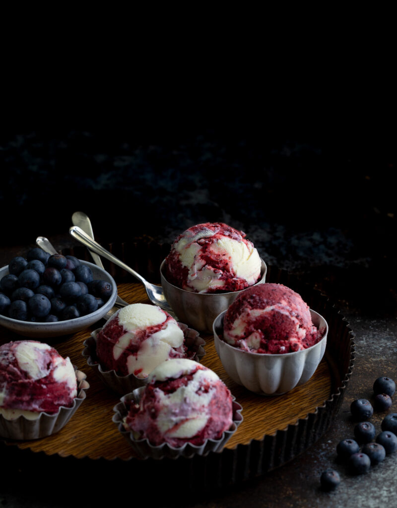 Scoops of blueberry sorbet vanilla swirl ice-cream in metal bowls on a serving tray with some fresh blueberries.