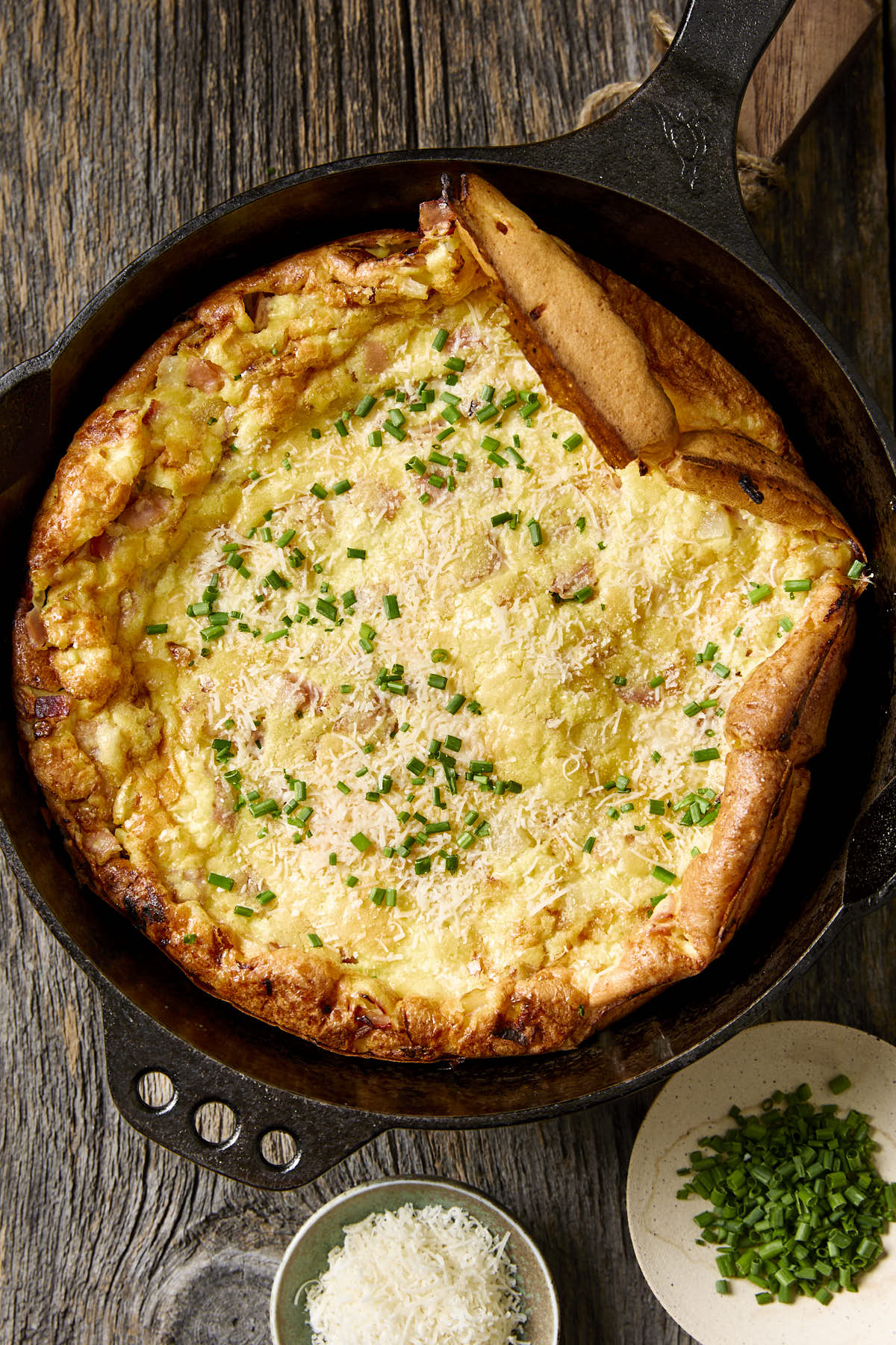 Savory dutch baby in a cast iron skillet next to some chopped chives and grated cheese.