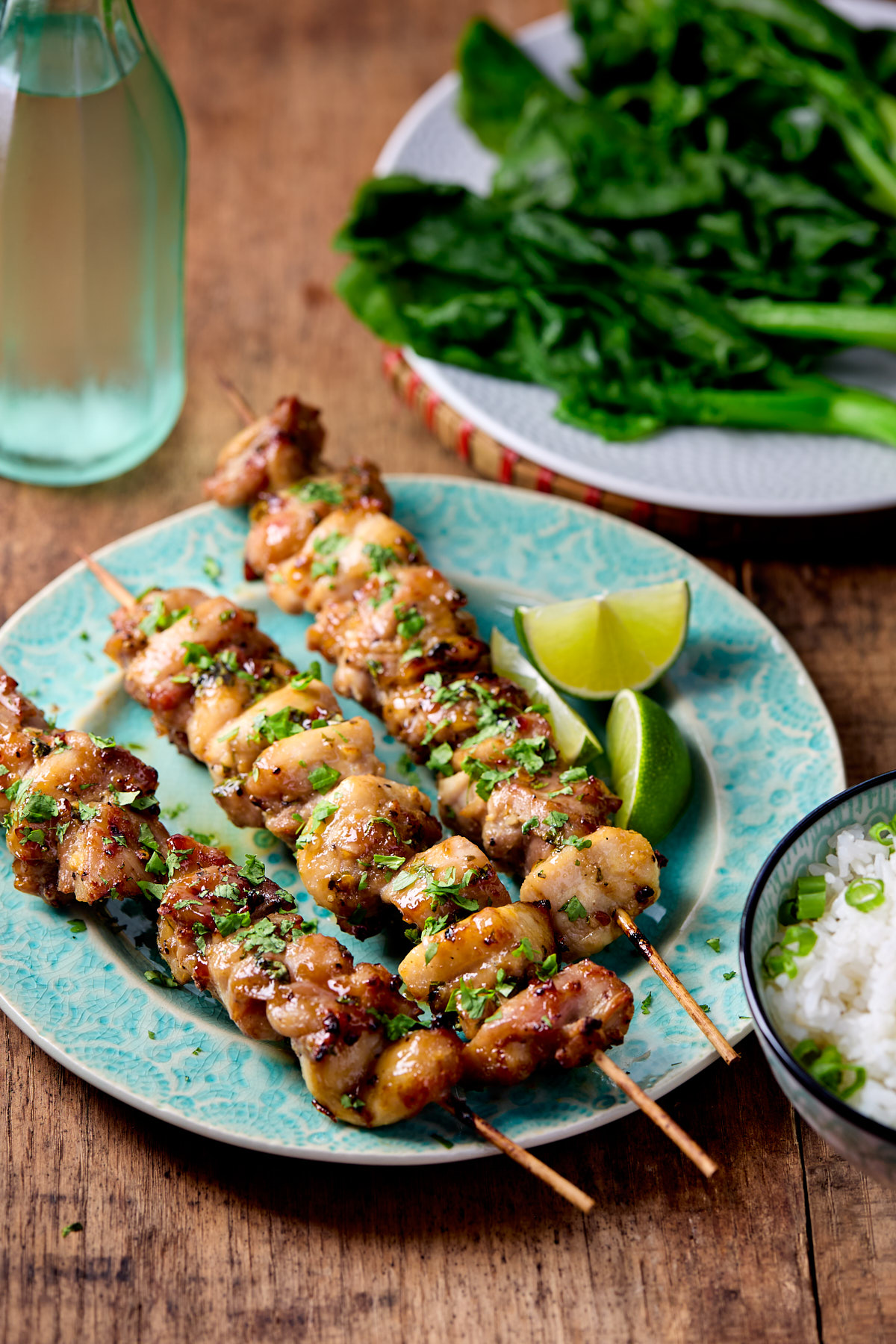 Three chicken skewers on a green plate next a plate of greens and a bowl of rice.
