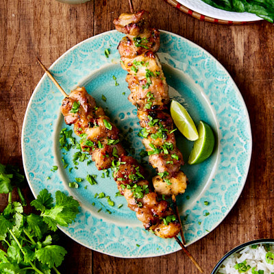 Two chicken skewers on a green plate next to a plate of greens and bowl of rice.