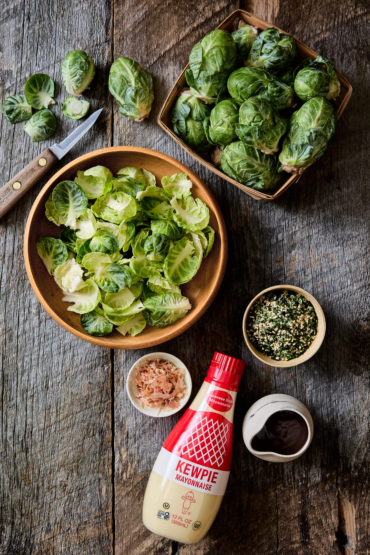 Fresh Brussels sprouts, a bottle of kewpie mayo, some bonito flakes, and some furikake on a wooden tabletop.