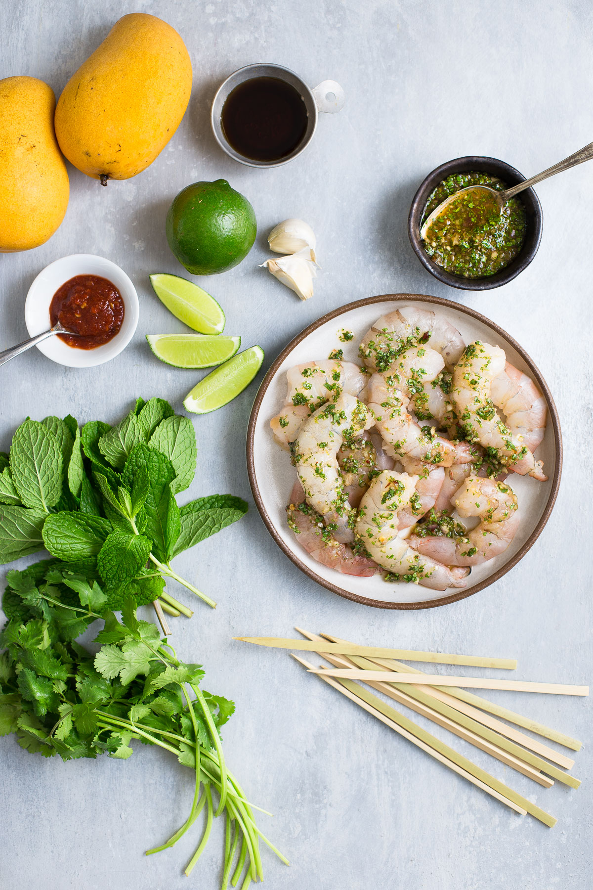 Some raw shrimp on a plate next to some mangos, limes, mint, cilantro, and garlic.
