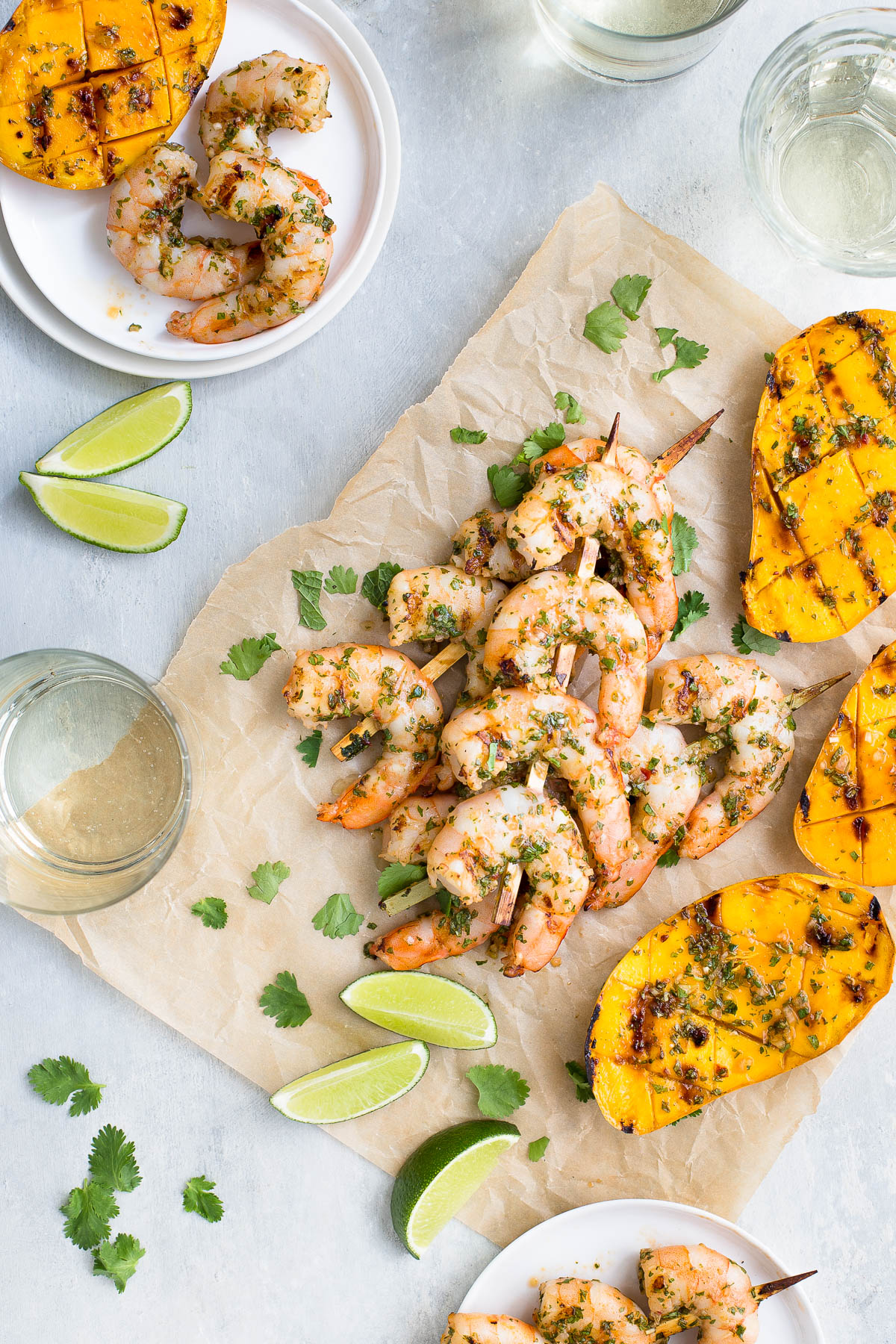 Skewers of grilled shrimp and grilled mango halves on parchment paper next to some glasses of wine and small white plates.