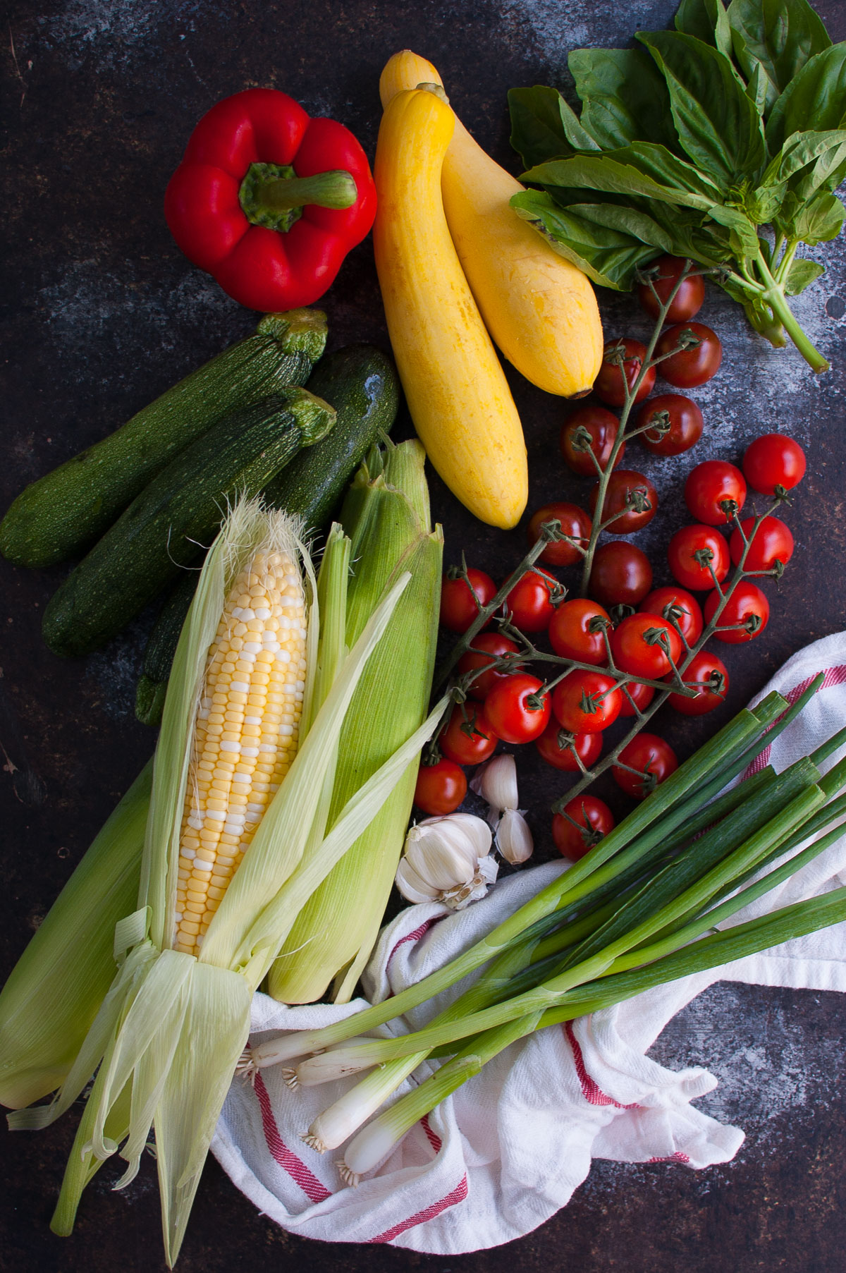 Some corn, green onions, squash, tomatoes, and a red bell pepper on a table top.