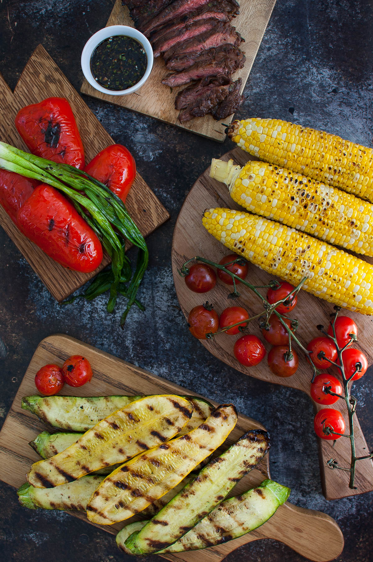 Some grilled steak, corn, squash, tomatoes, and bell peppers on various wooden boards.