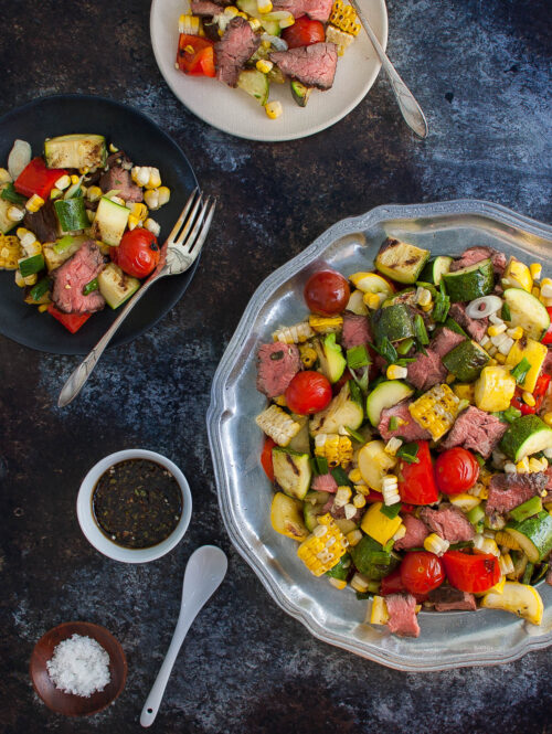 Grilled steak and vegetables salad on a platter next to small salad plates.
