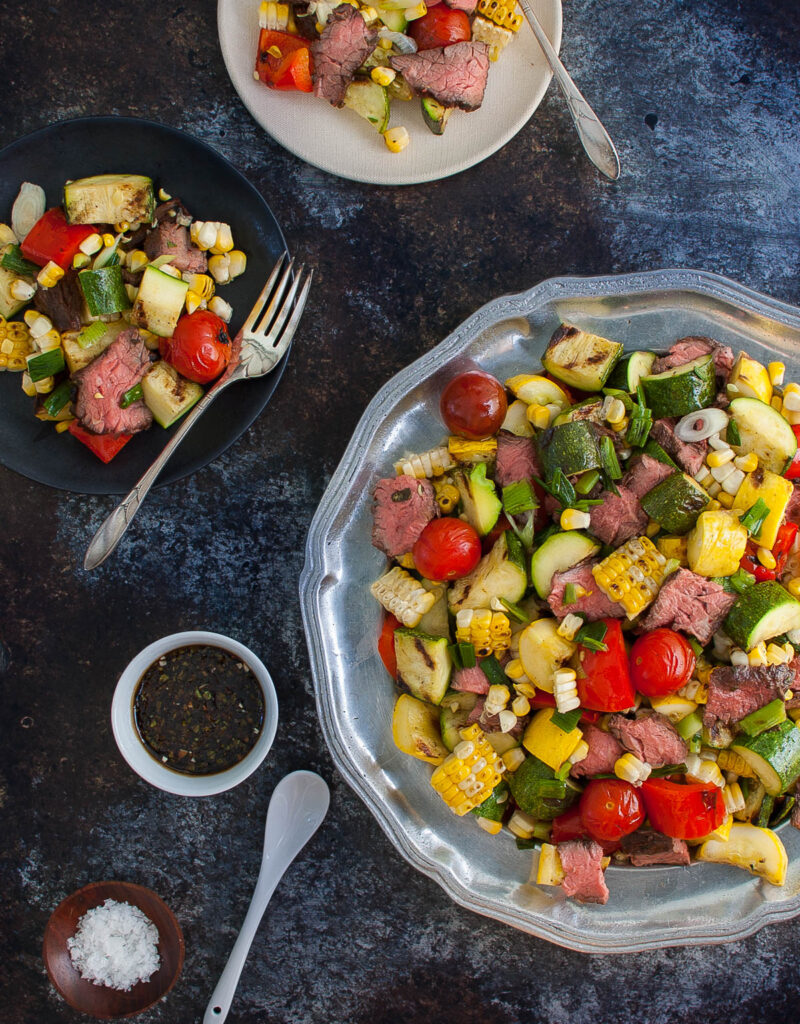 Grilled steak and vegetables salad on a platter next to small salad plates.