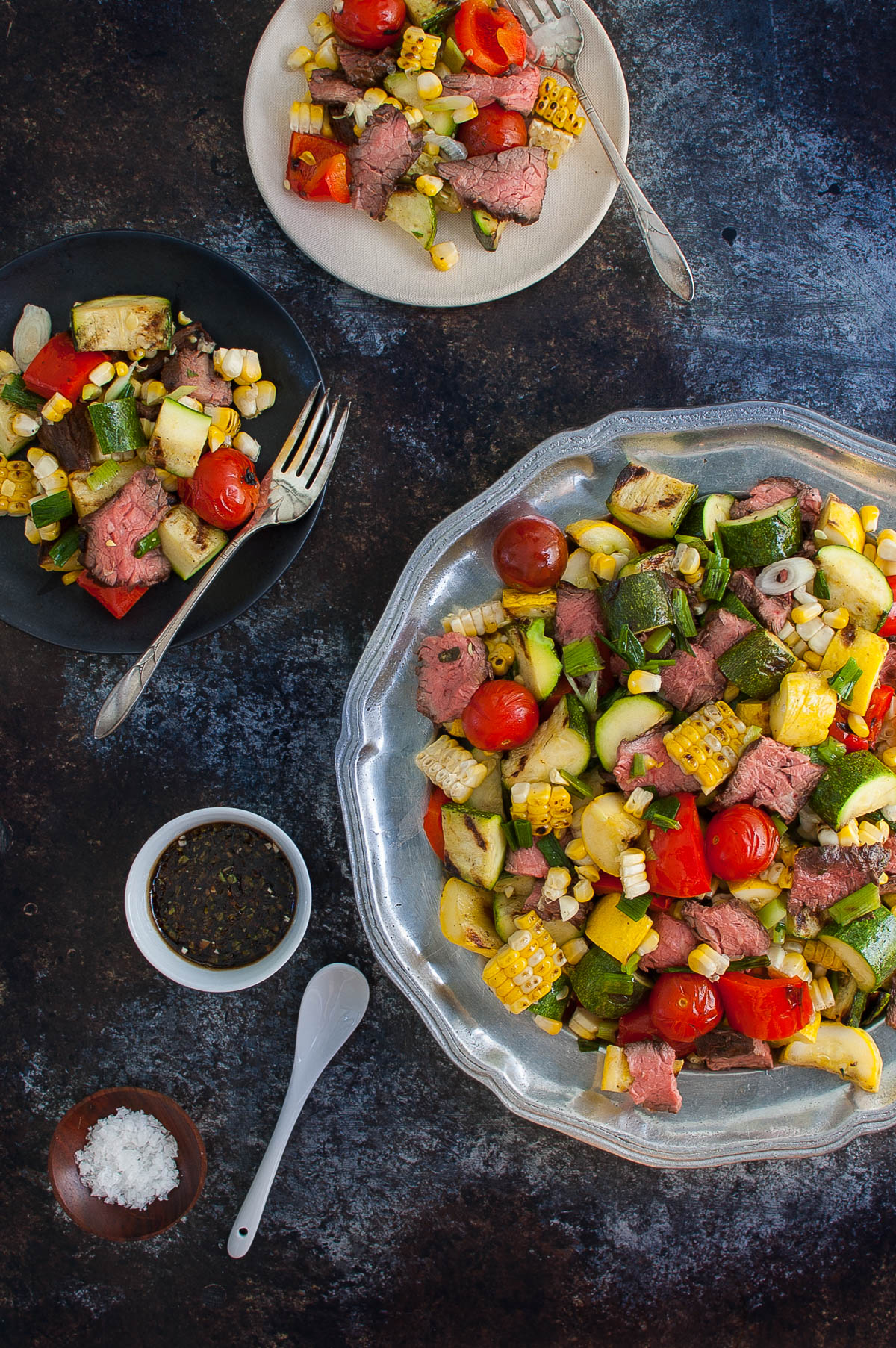 Grilled steak and vegetables salad on a platter next to small salad plates.