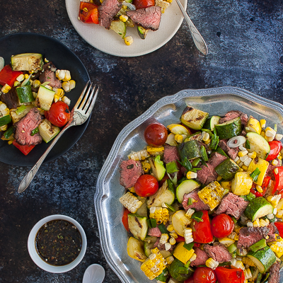 Grilled steak and vegetables salad on a platter next to small salad plates.