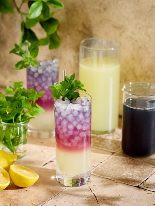Glasses of blackberry tea lemonade next to some lemons, mint sprigs, and two pitchers.