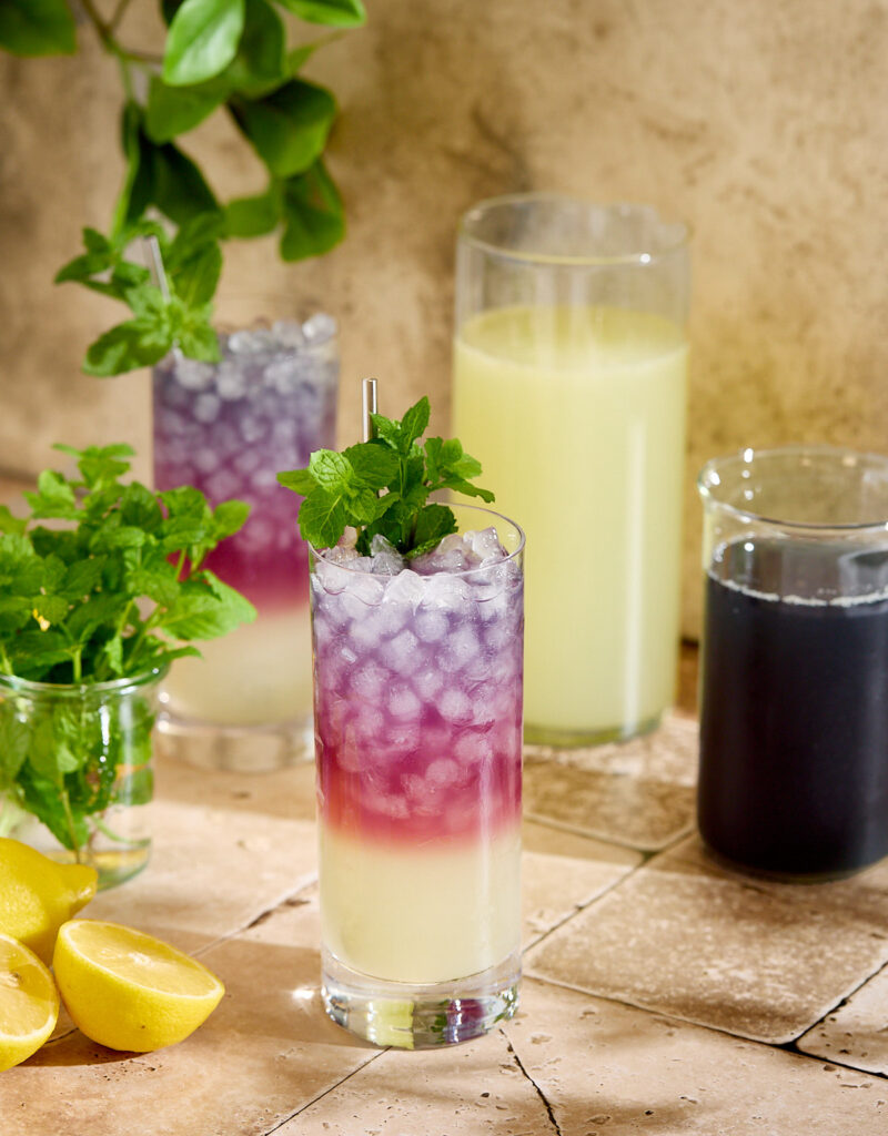 Glasses of blackberry tea lemonade next to some lemons, mint sprigs, and two pitchers.