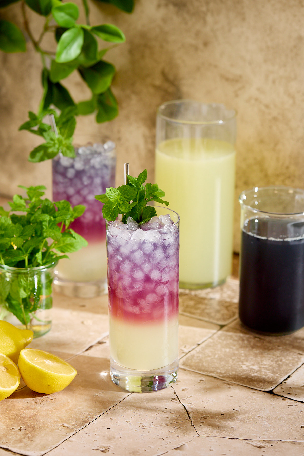 Glasses of blackberry tea lemonade next to some lemons, mint sprigs, and two pitchers.