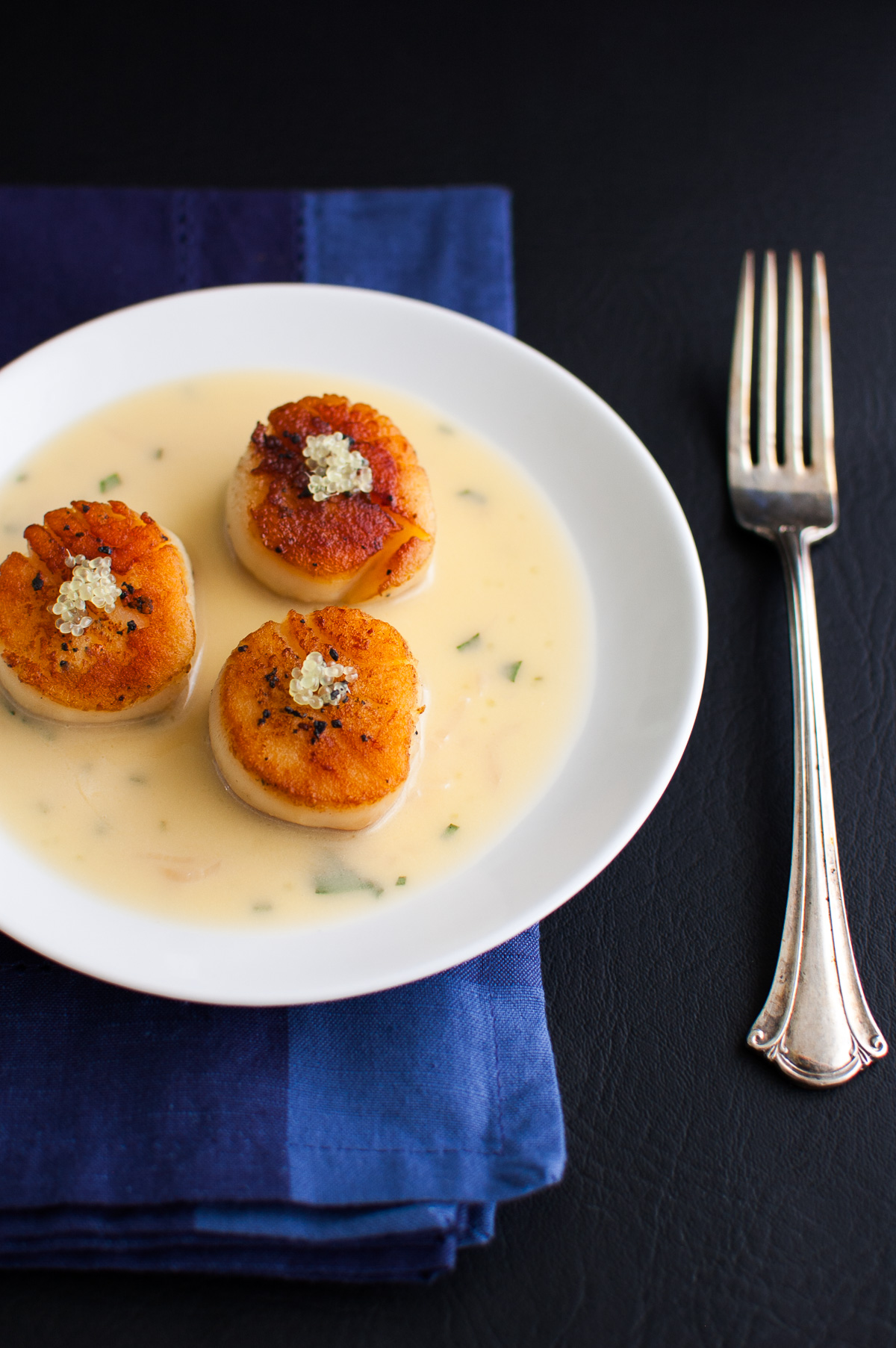 Three scallops with finger lime beurre blanc on a white plate next to a fork and blue napkin.